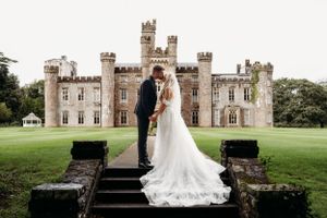 A couple poses on stone steps in front of a majestic castle with turrets and towers on a sprawling green lawn.