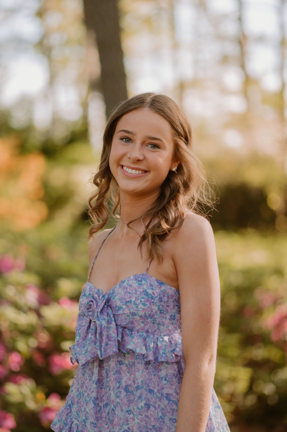 Young woman in a floral dress smiles in a sunlit garden with colorful flowers and trees in the background.