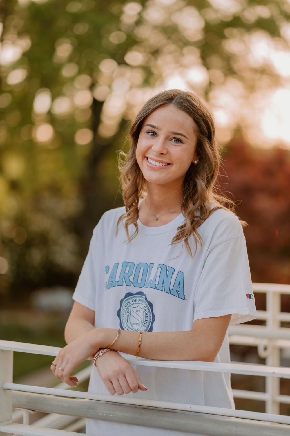 Smiling young woman with long hair in a white "Carolina" shirt, leaning on a white railing, with greenery in the backgro