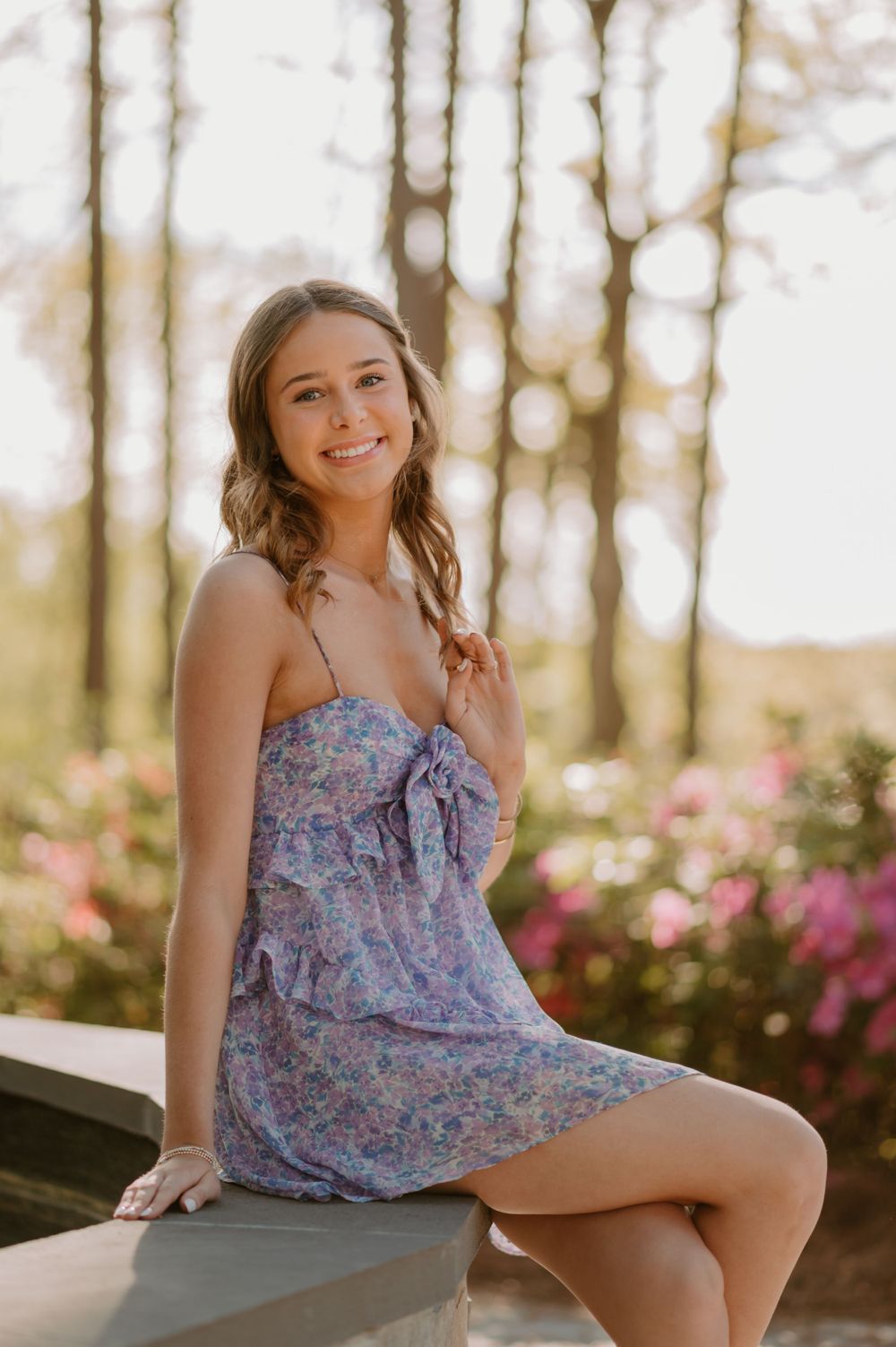 A woman in a floral dress sitting on a bench outdoors, smiling with a background of trees and blooming flowers.