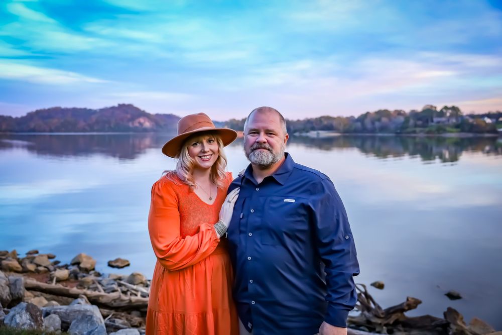 A couple poses by a serene lake with mountains in the background during sunset.