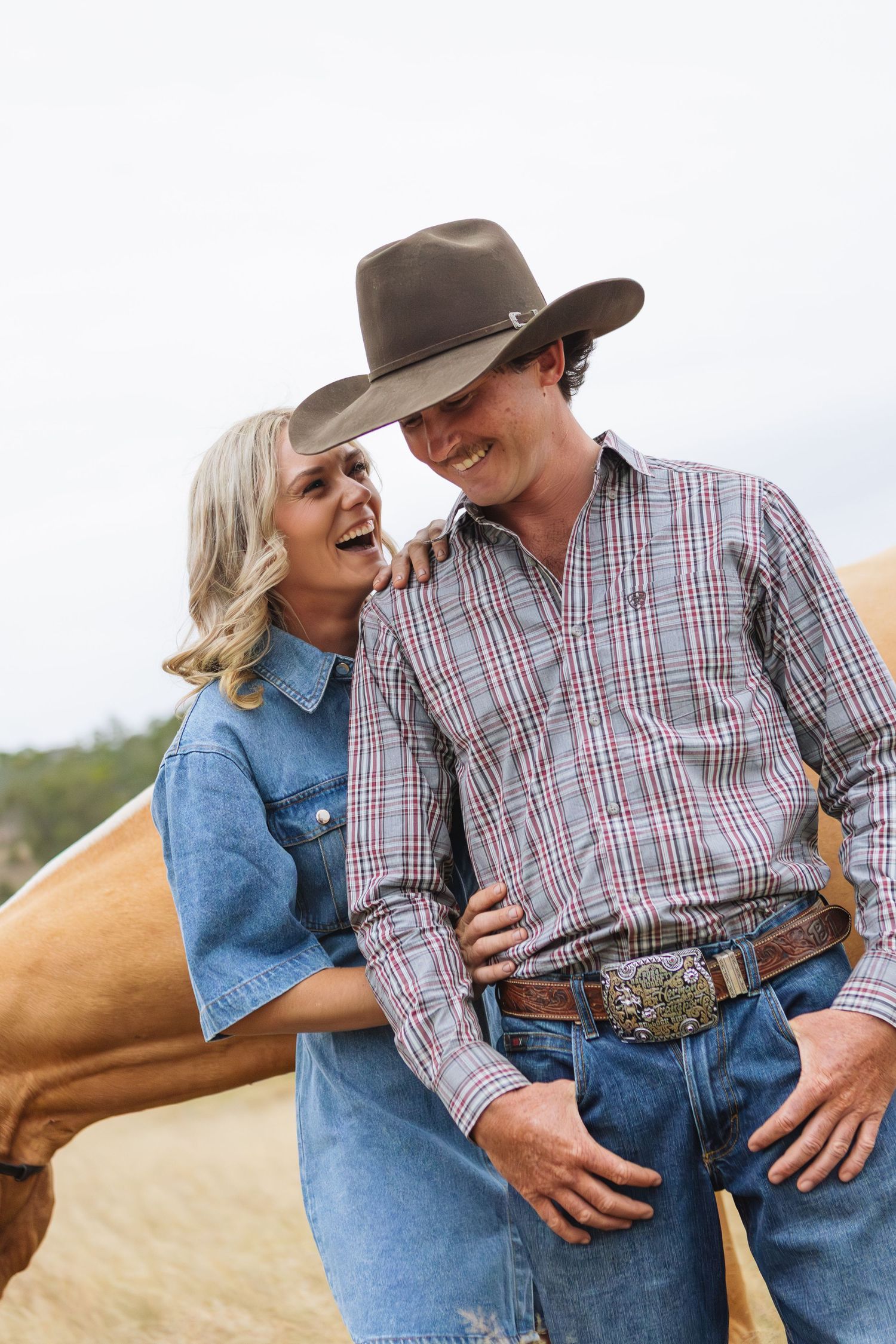 girl in cowgirl boots and denim dress hugs man in cowboy hat during engagement photoshoot with horse in the background