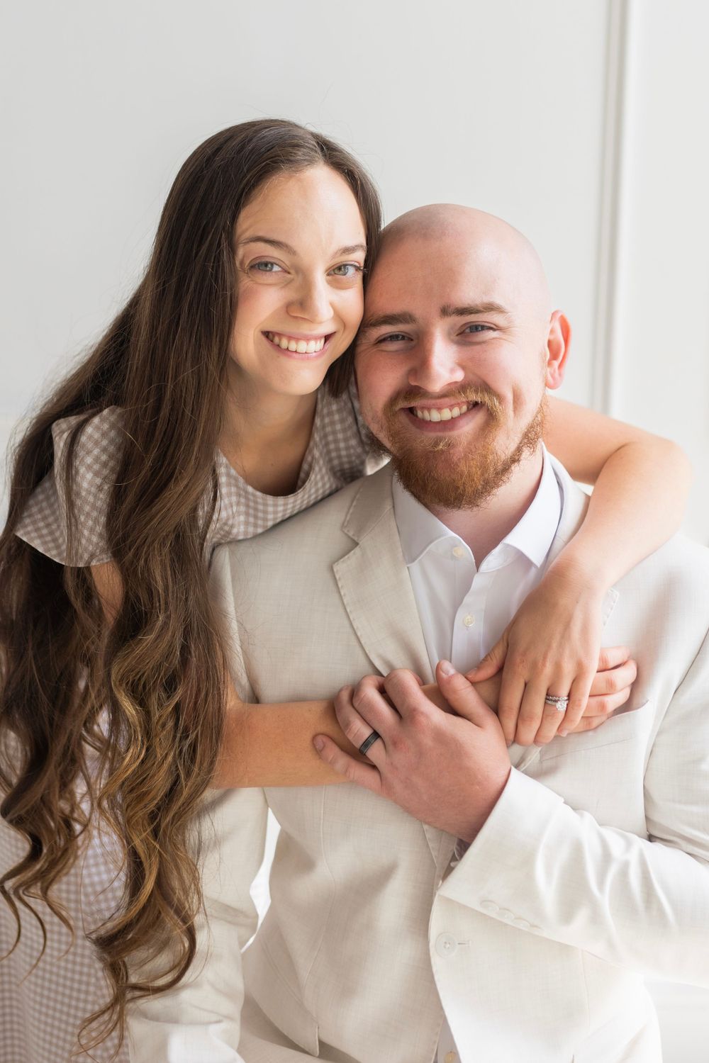 A couple in white and beige outfits embrace and smile warmly at the camera while posing for a romantic portrait.