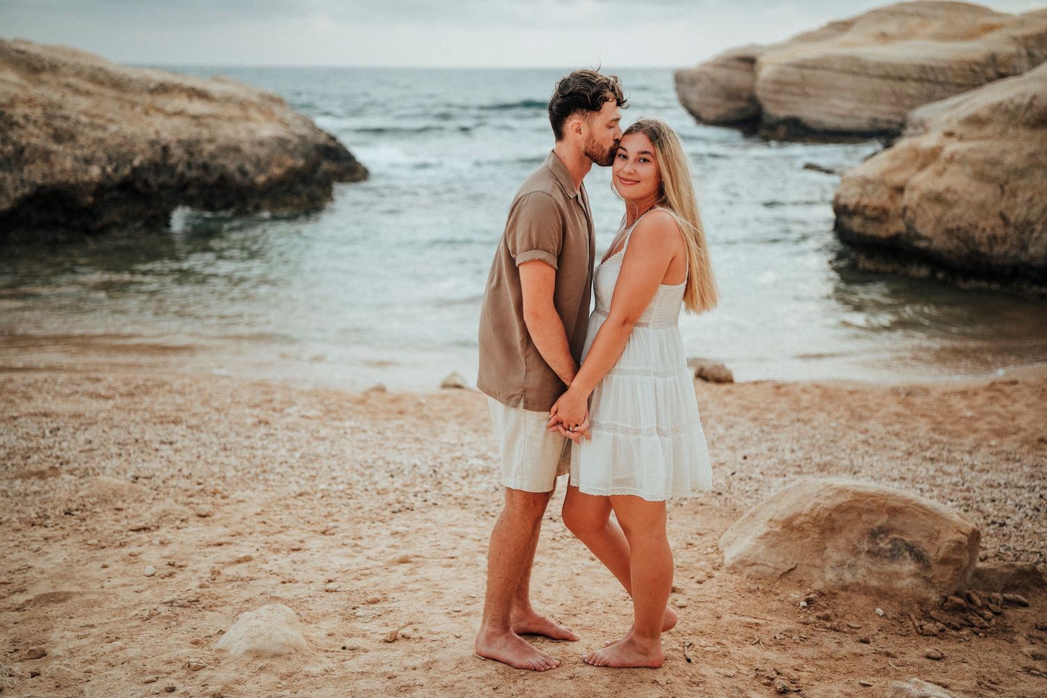 A couple shares an intimate moment on a sandy beach surrounded by rocky coastal formations at sunset.