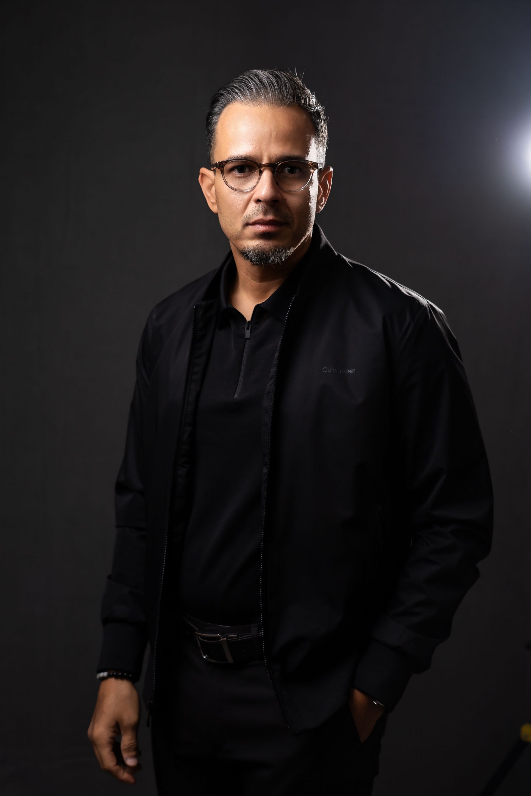 Man in brown leather jacket and tan pants smiling against white studio background.