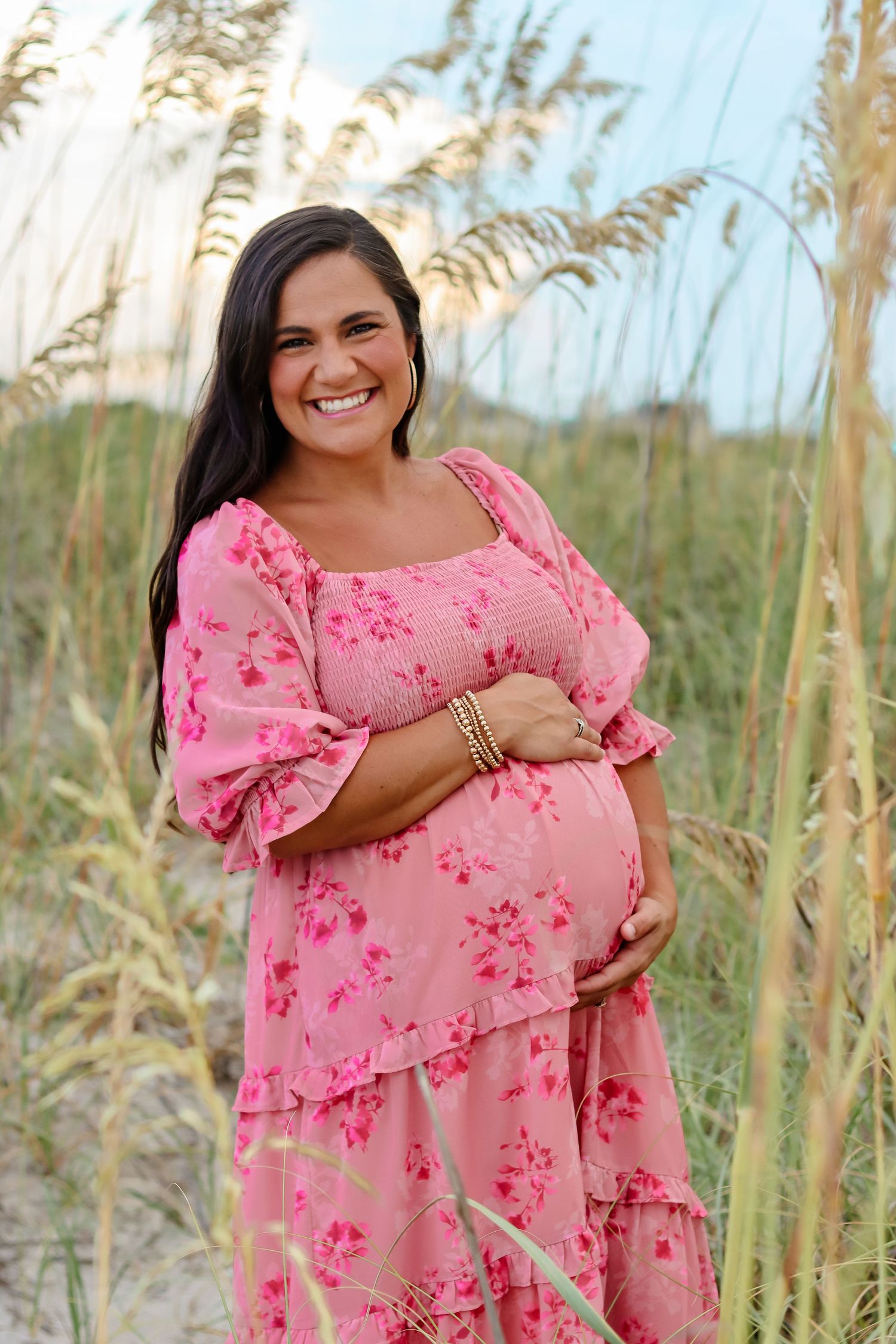 A maternity portrait of an expecting mother in a pink floral dress among sea oats.
