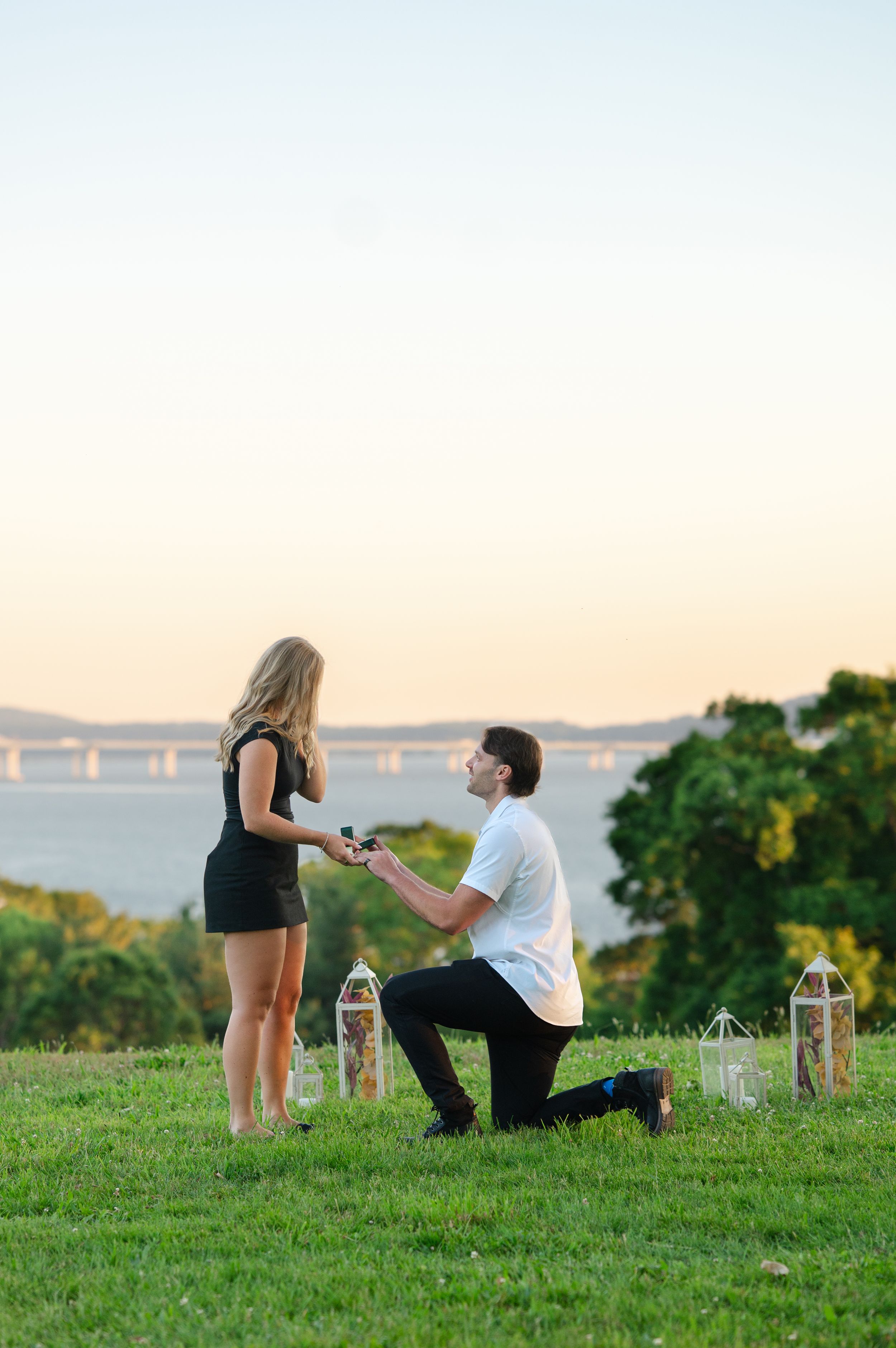 Man proposes on one knee to woman in black dress at sunset overlooking a scenic waterway with lanterns nearby.