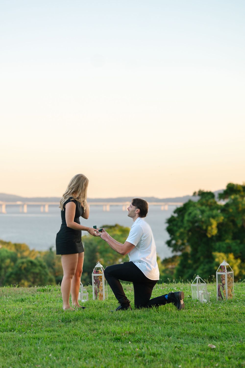 Man proposes on one knee to woman in black dress at sunset overlooking a scenic waterway with lanterns nearby.