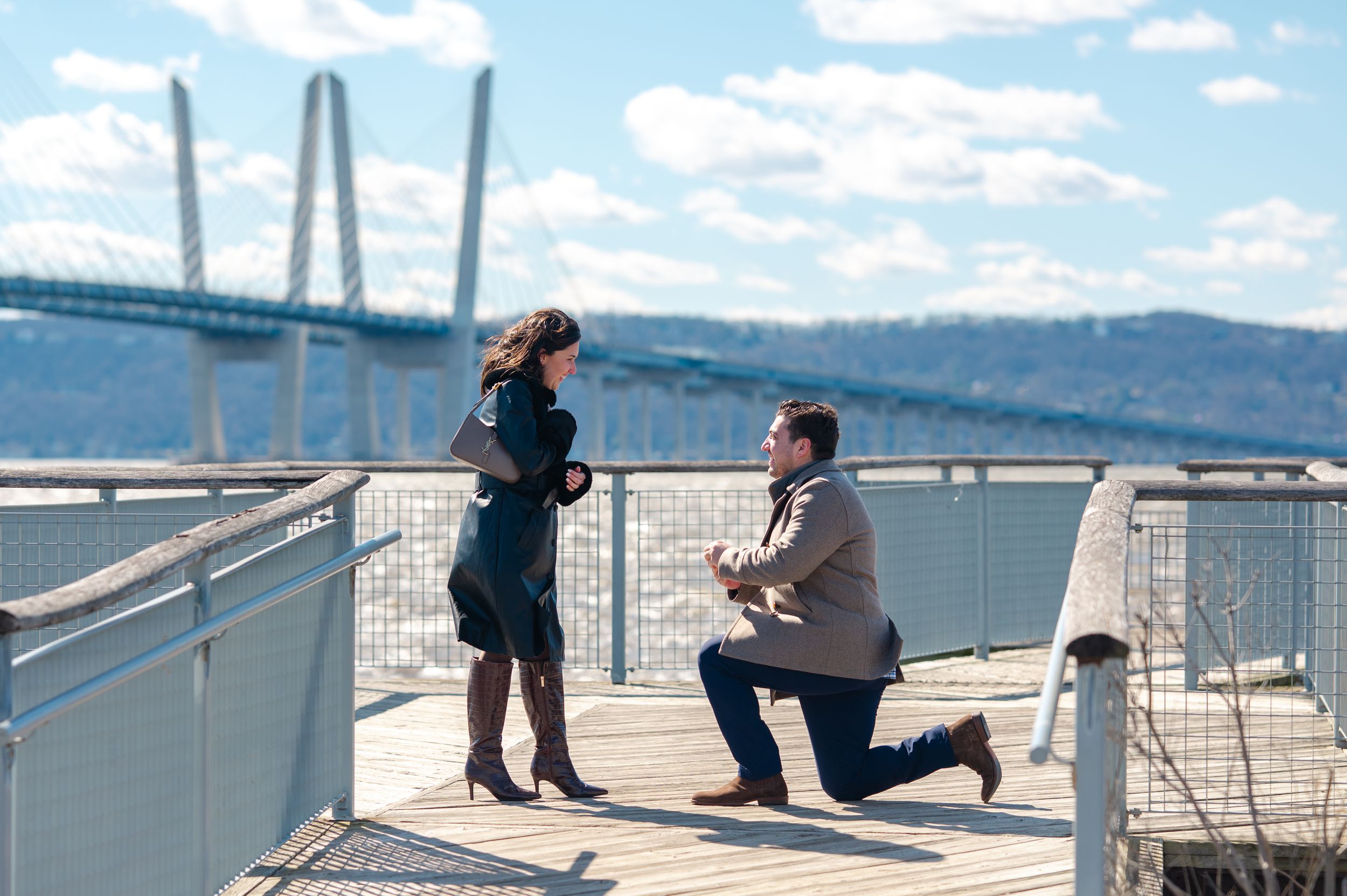 Man proposes to woman on a waterfront deck with the Tappan Zee Bridge in the background on a sunny day.