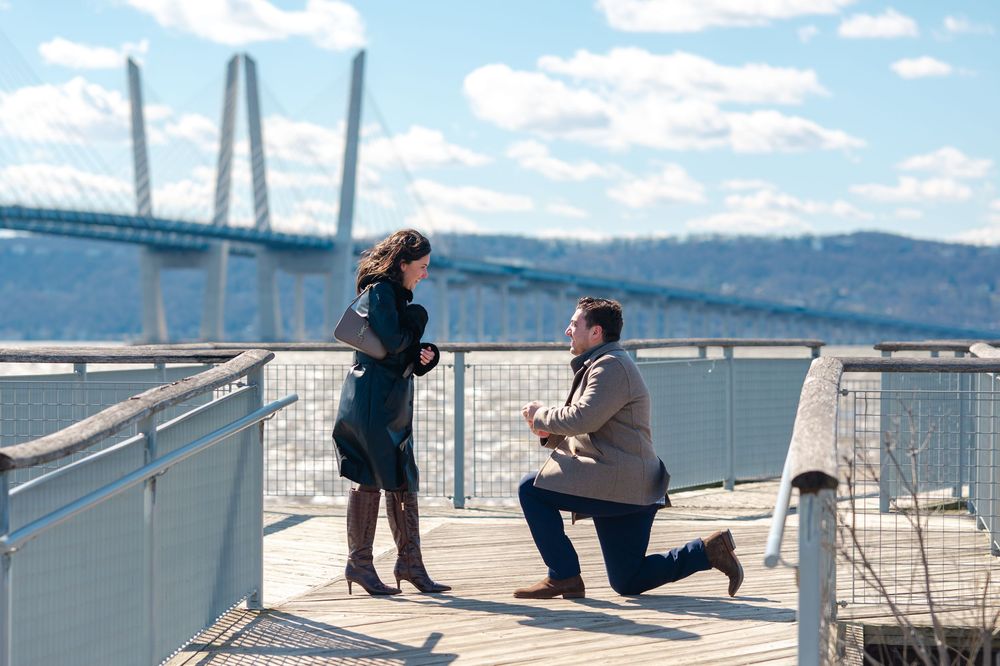 Man proposes to woman on a waterfront deck with the Tappan Zee Bridge in the background on a sunny day.