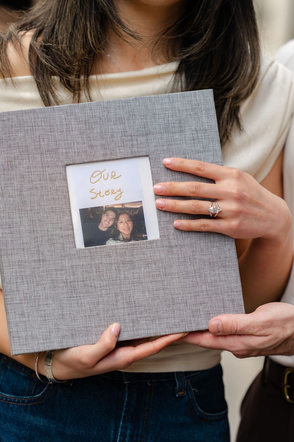 Couple holding a gray linen photo album with a Polaroid picture and "Our Story" text on the cover.