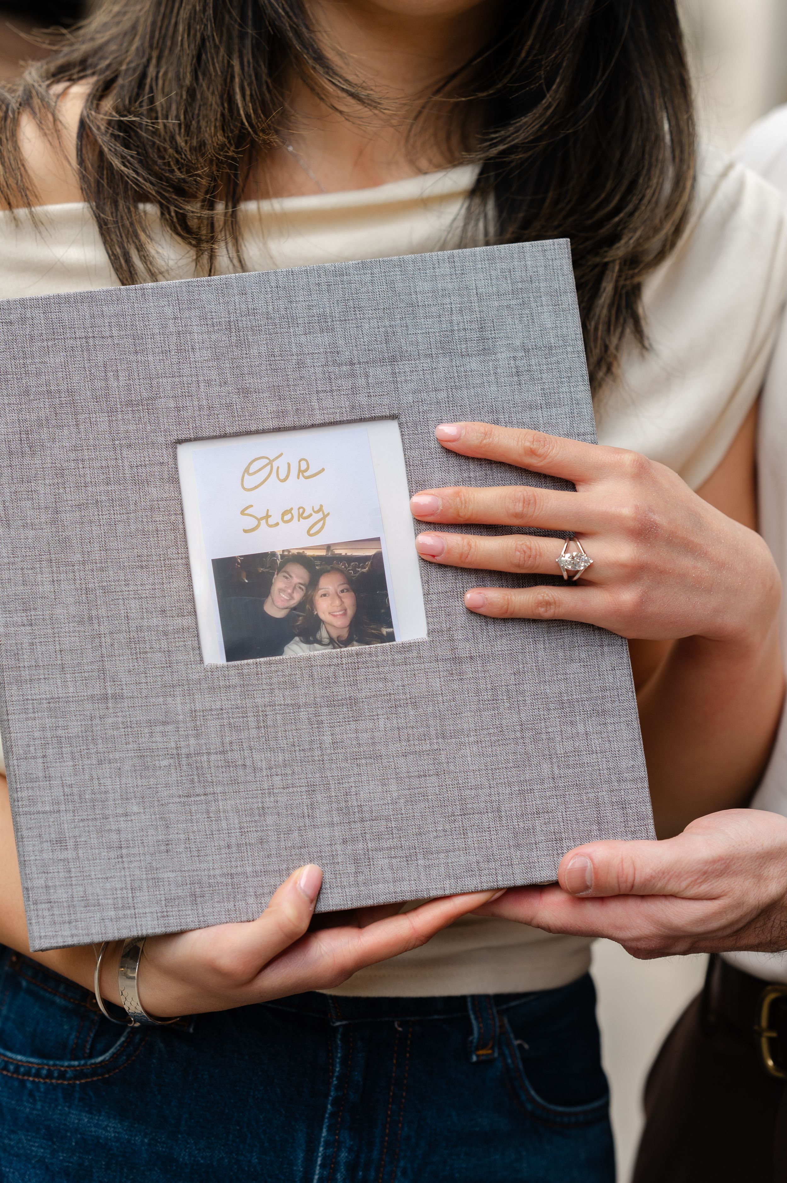 Couple holding a gray linen photo album with a Polaroid picture and "Our Story" text on the cover.