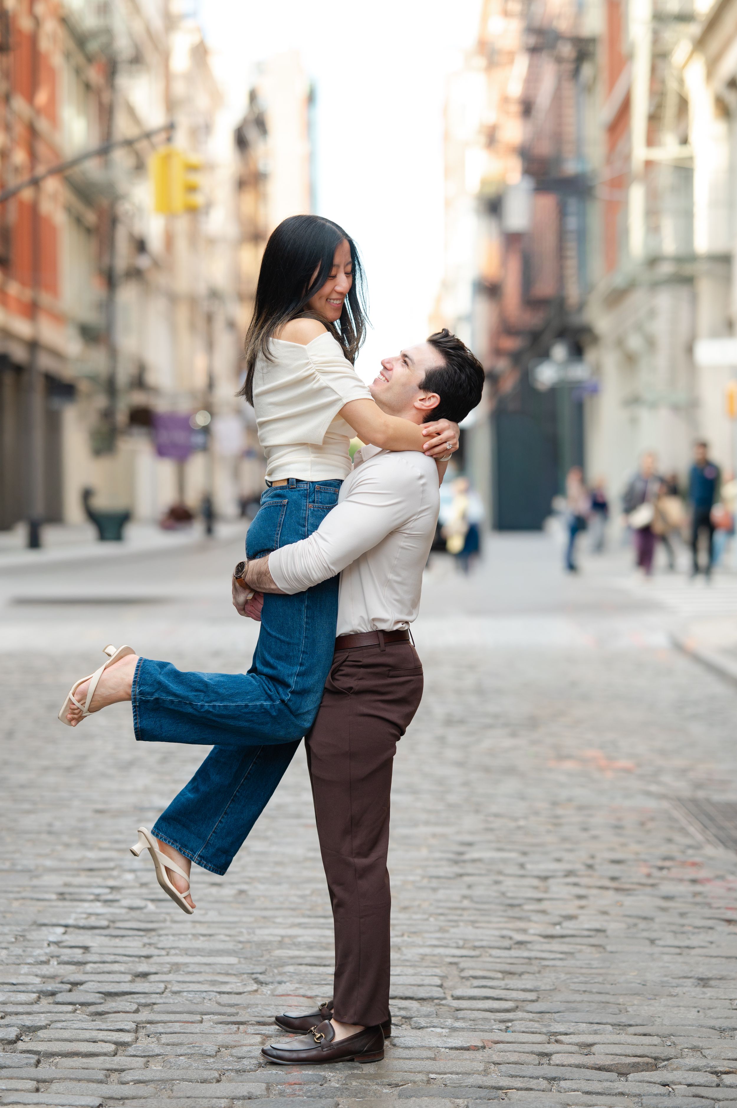 A man lifts a smiling woman on a cobblestone city street, both dressed in casual chic outfits.