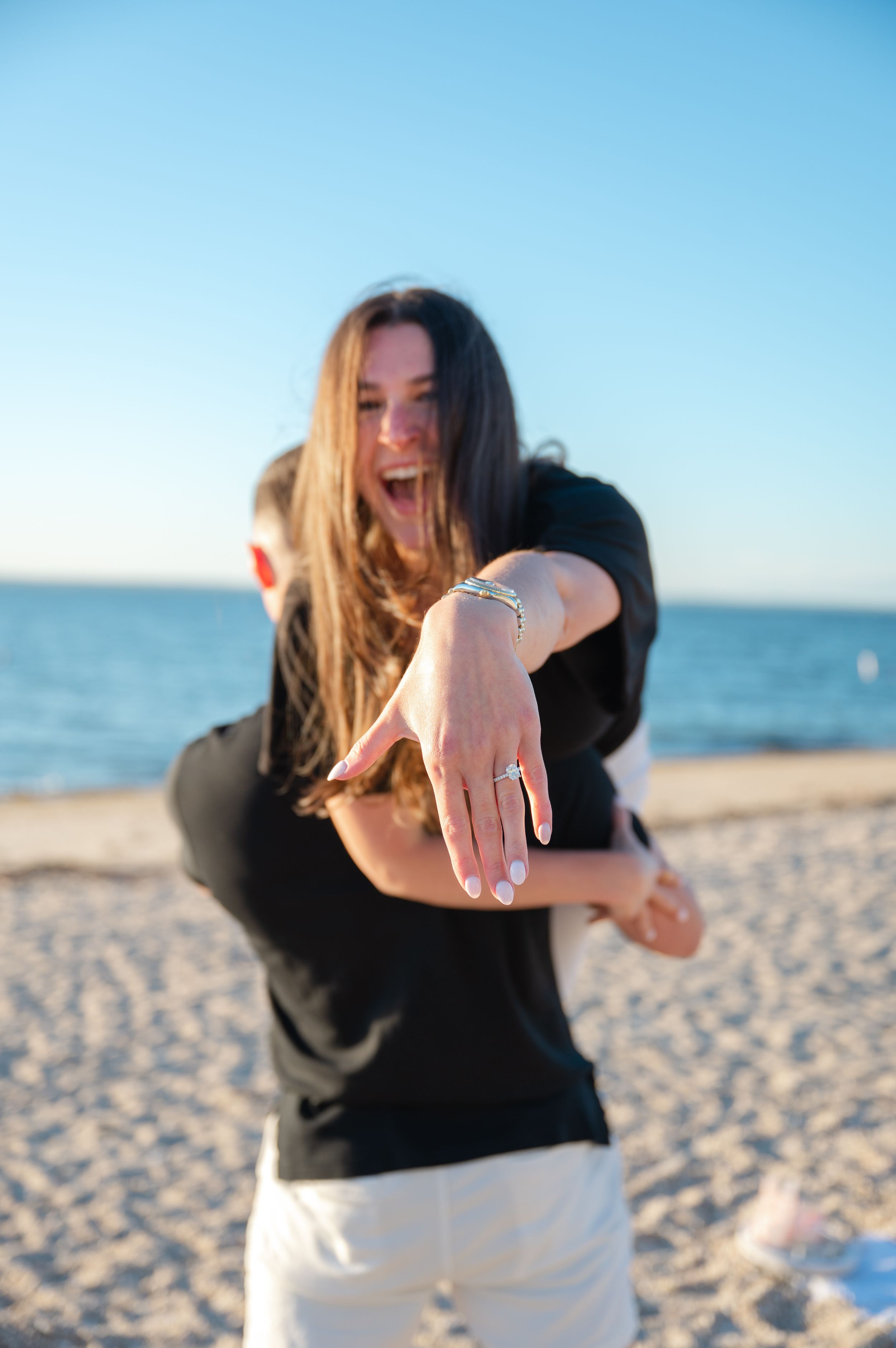 Happy woman showing off engagement ring on beach, embraced by partner, with ocean and clear blue sky in background.