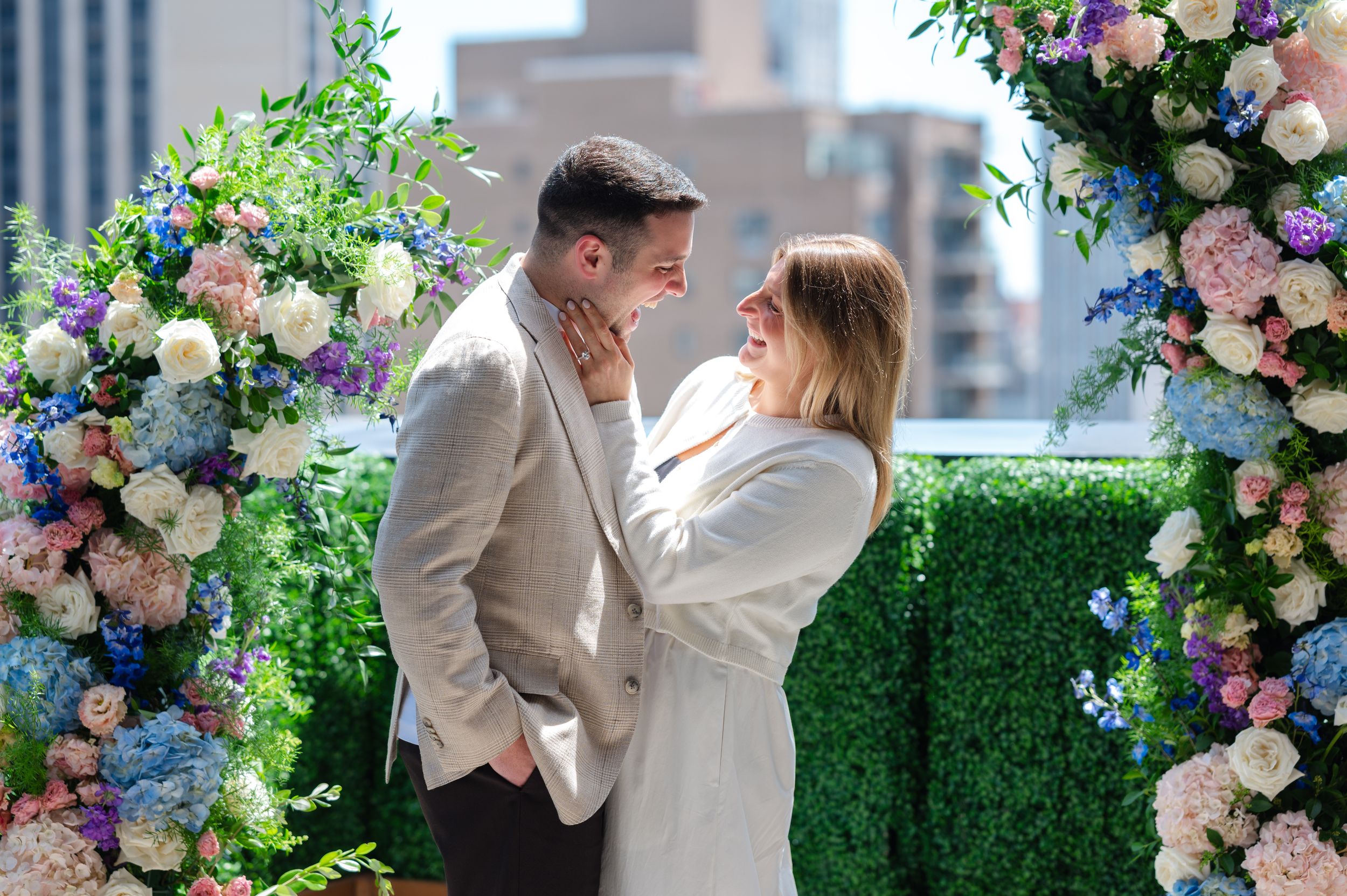 Couple sharing a romantic moment under a floral arch with pink and blue flowers on a rooftop.