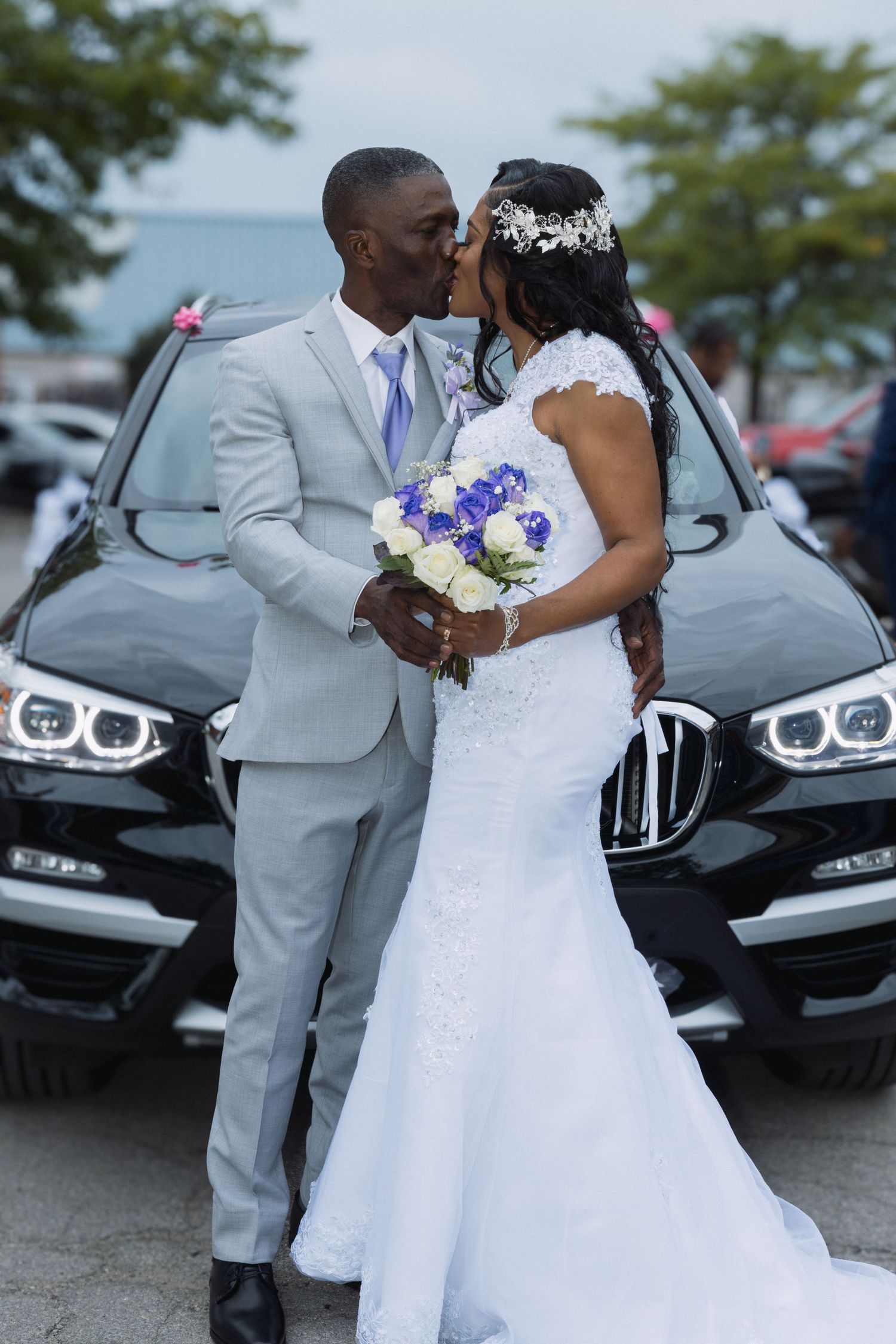 Newlyweds kiss in front of black BMW SUV, bride holding blue and white bouquet in wedding attire. Wedding taken by BEXPRO in Vaughan, ON.