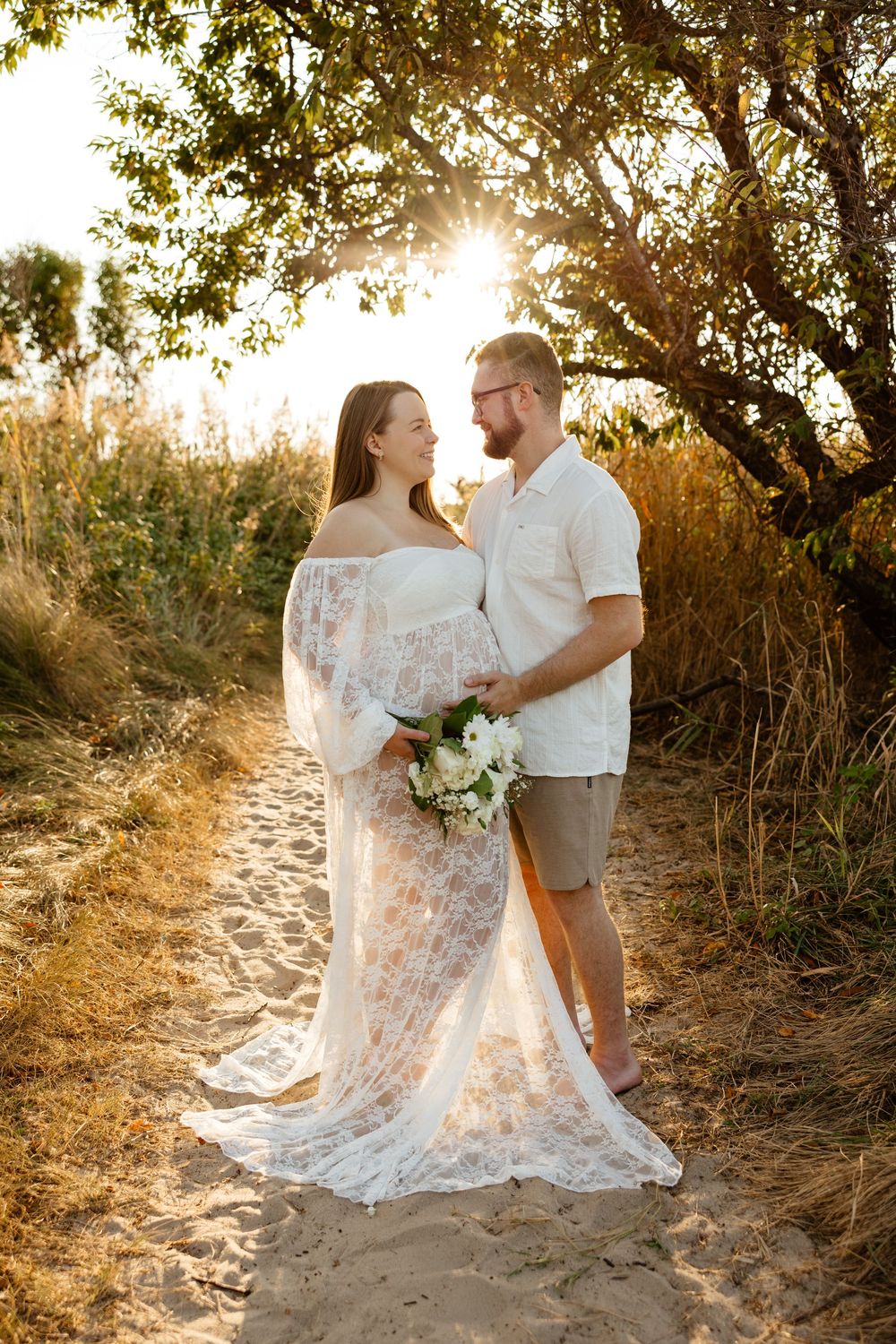A pregnant couple shares a romantic moment at sunset holding a white floral bouquet in Cape May during a maternity session.