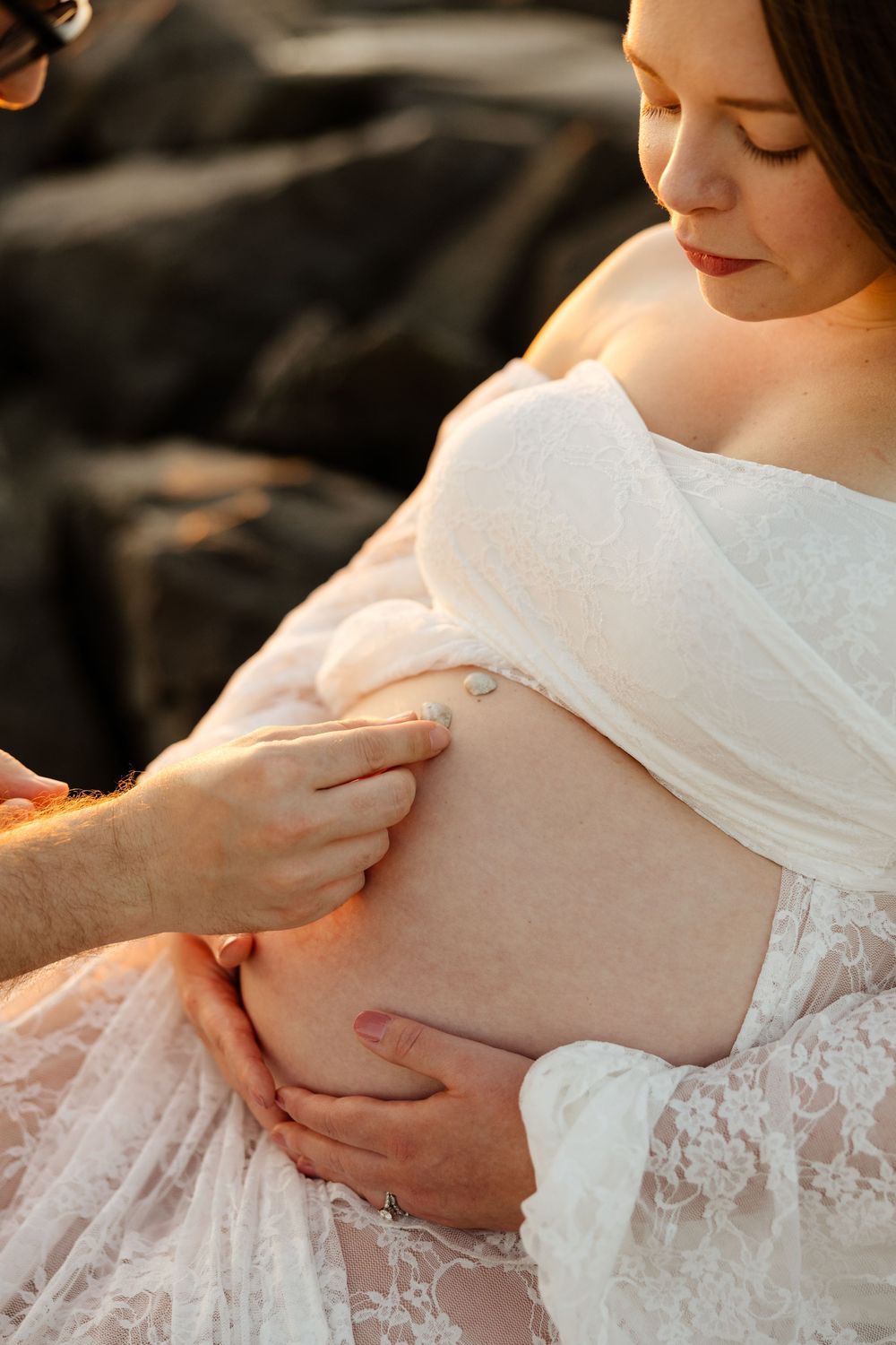 Hands gently touching a pregnant woman's belly while she wears a white lace maternity dress in warm sunlight.