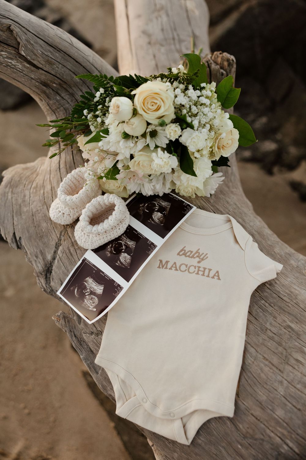 A collection of cream colored shoes and baby onesies arranged with white floral bouquets on weathered wood.