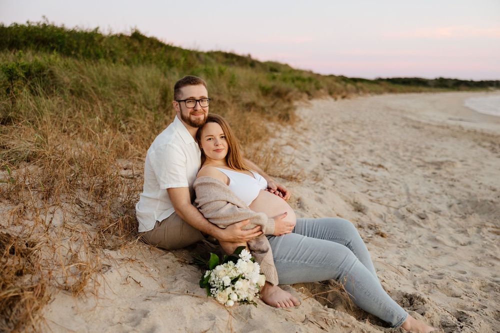 A couple relaxes together on a sandy beach at sunset with a white floral bouquet lying beside them.