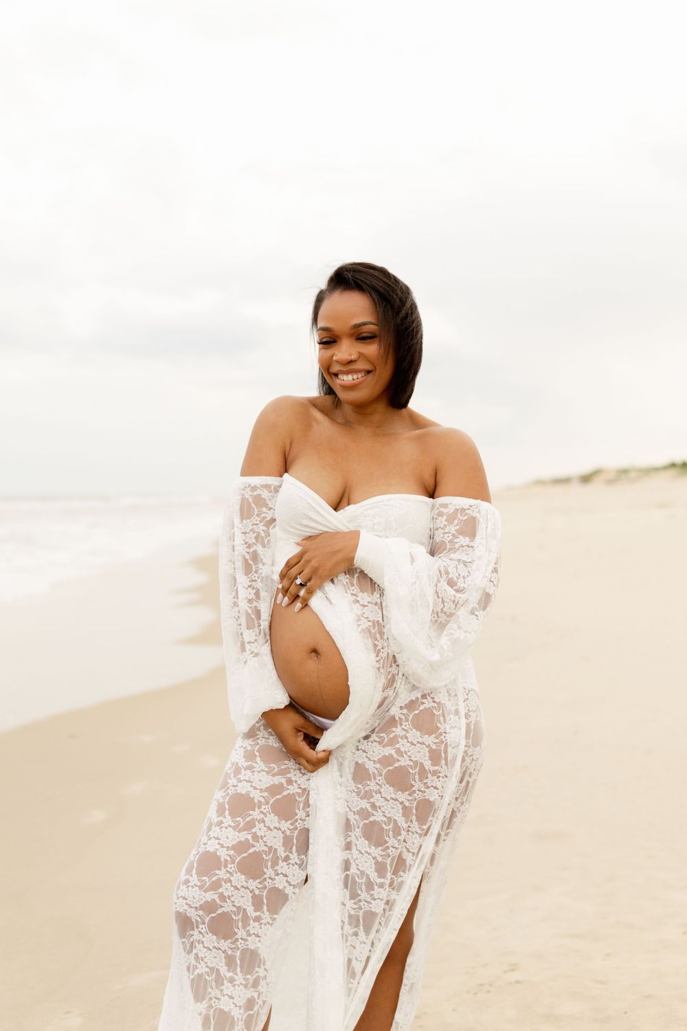 Beautiful maternity portrait on a beach with white lace dress and gentle smile, showcasing pregnancy joy at sunset.