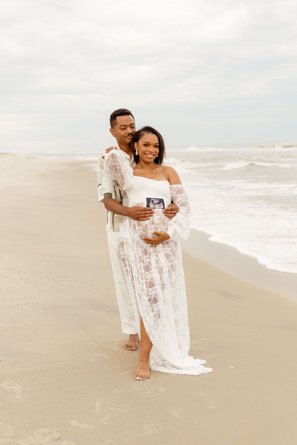 Couple in white attire standing on beach holding ultrasound photo during pregnancy announcement photoshoot.