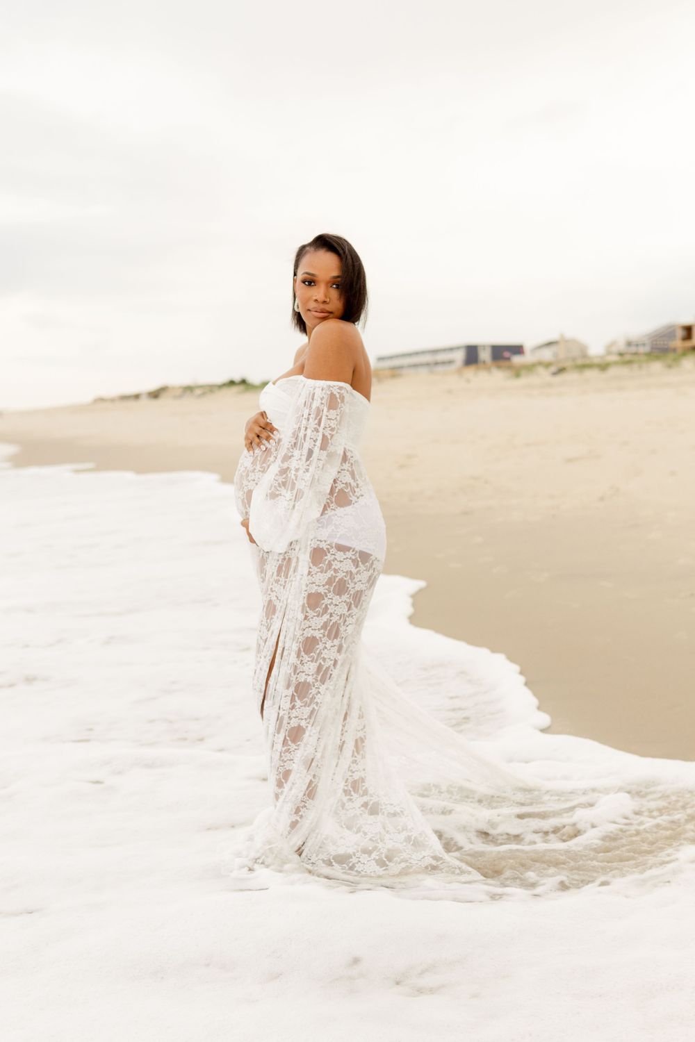 A graceful figure in a flowing white lace dress poses elegantly on a sandy beach at sunset.