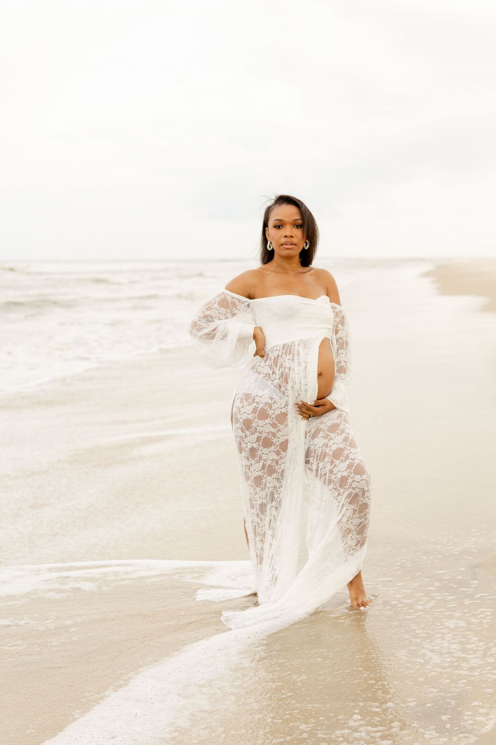A person in a flowing white lace off-shoulder dress poses elegantly on a misty beach at sunrise.