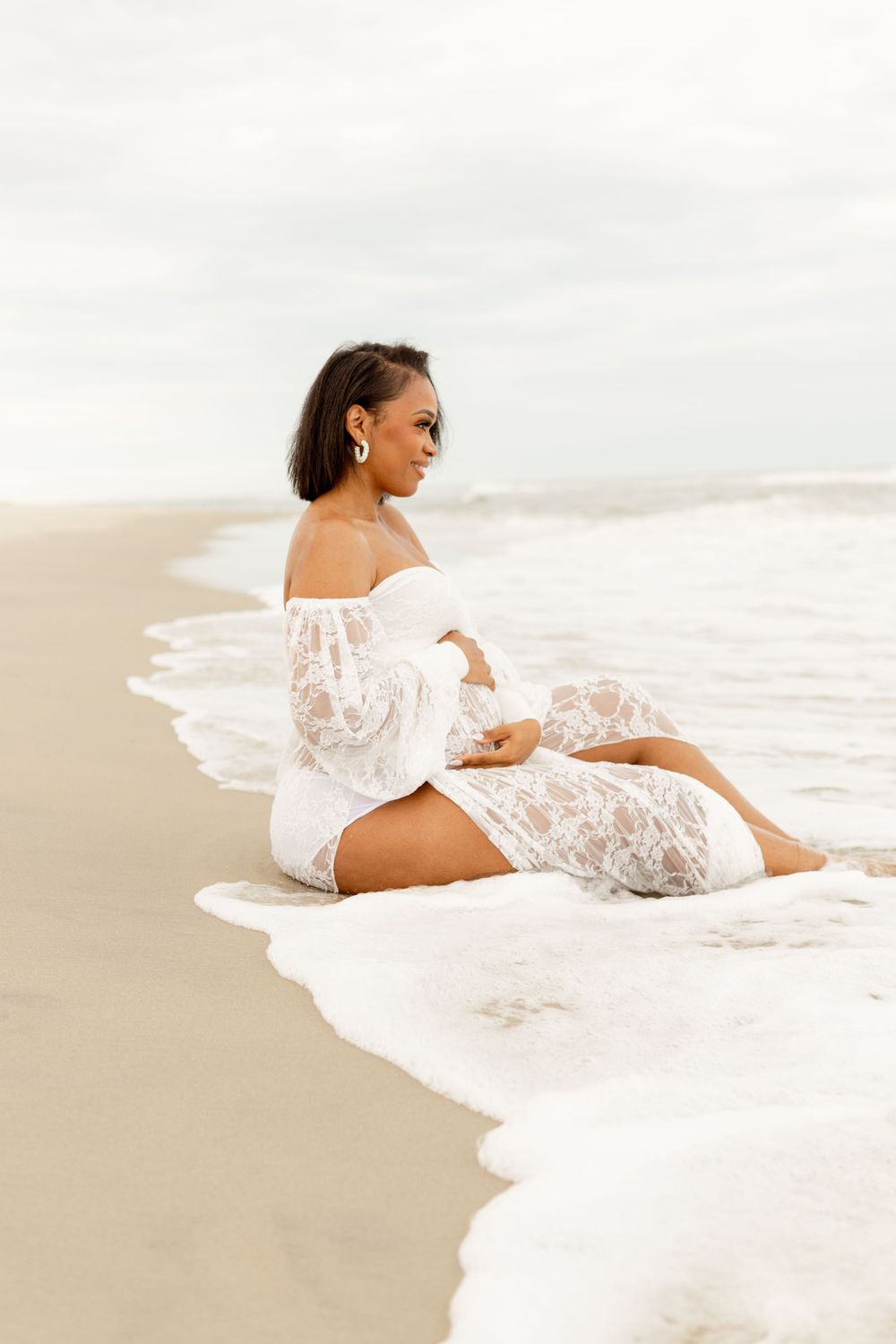 Woman in ethereal white lace dress sits peacefully at the edge of ocean waves on a sandy beach at sunset.