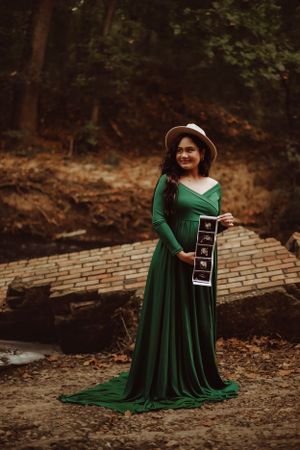 Woman in emerald green dress holding ultrasound strip in wooded setting.