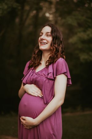 Expectant mother in a pink dress laughs joyfully while cradling her baby bump in an outdoor setting.
