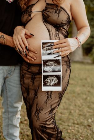 A couple holds ultrasound photos against a pregnant belly in a black lace maternity dress.
