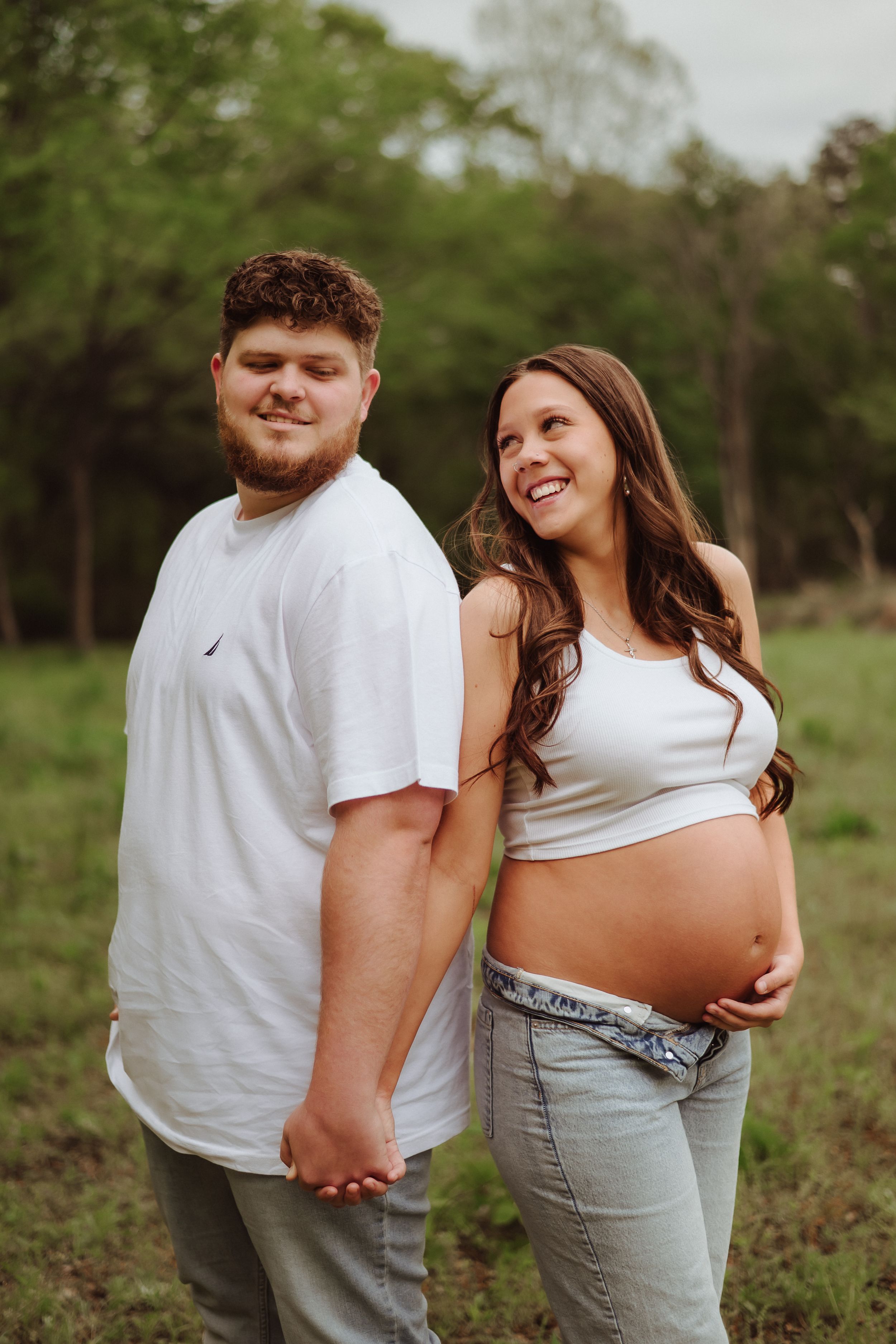 A couple poses together in casual attire for their maternity photos in a grassy field.