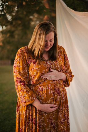 Pregnant woman in mustard floral dress holding belly during sunset maternity photo session.