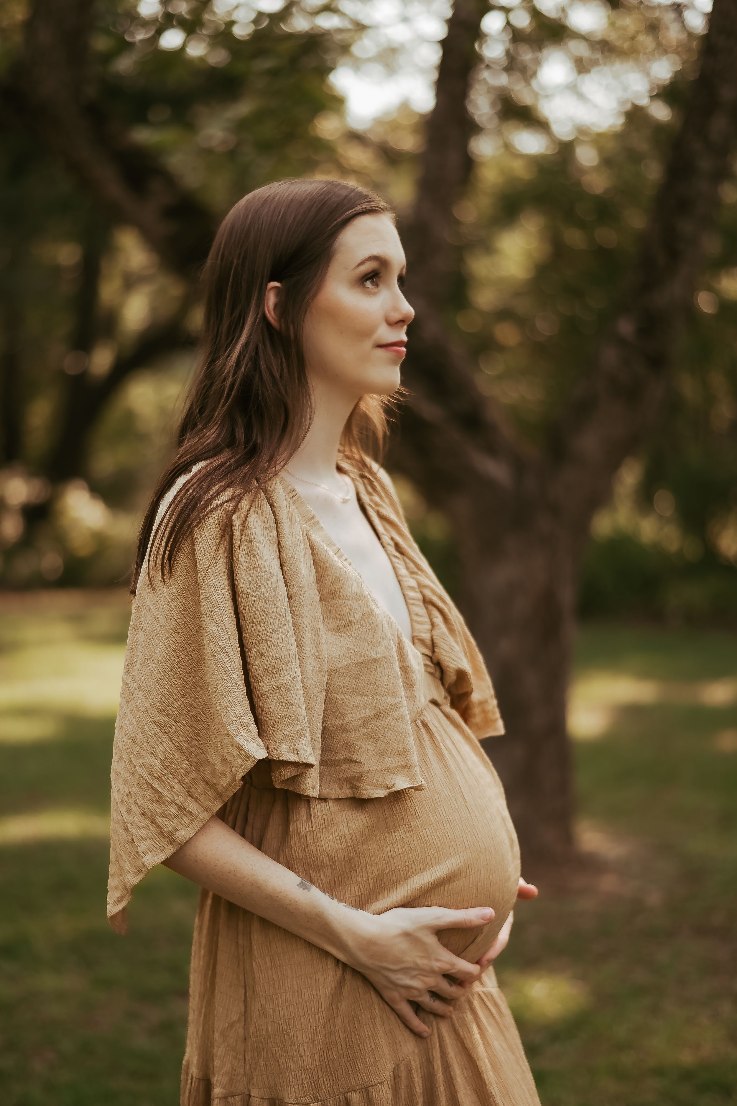A pregnant woman in a flowing beige dress poses in profile against a wooded backdrop.