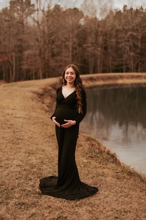 Maternity photo of woman in elegant black dress standing beside a serene lake at sunset.