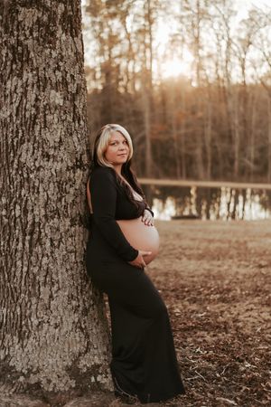 Maternity portrait of woman in black dress leaning against tree trunk at golden hour.