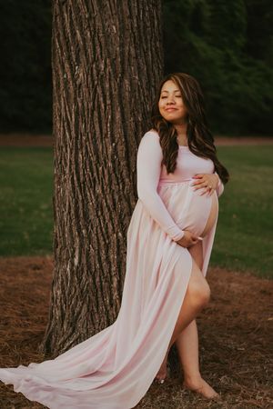 Expectant mother in flowing pink dress poses elegantly against a large tree trunk.