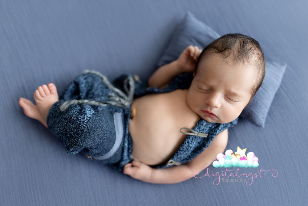 Sleeping newborn wearing blue outfit during professional photo session on gray backdrop.