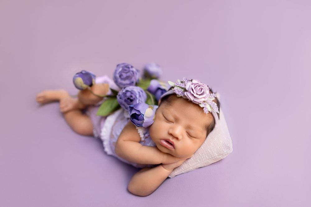 Newborn baby sleeping peacefully with purple floral accessories on lavender backdrop.