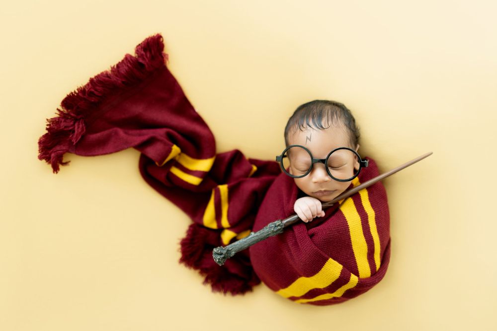 Harry Potter themed newborn photo with Gryffindor scarf and round glasses on cream background.