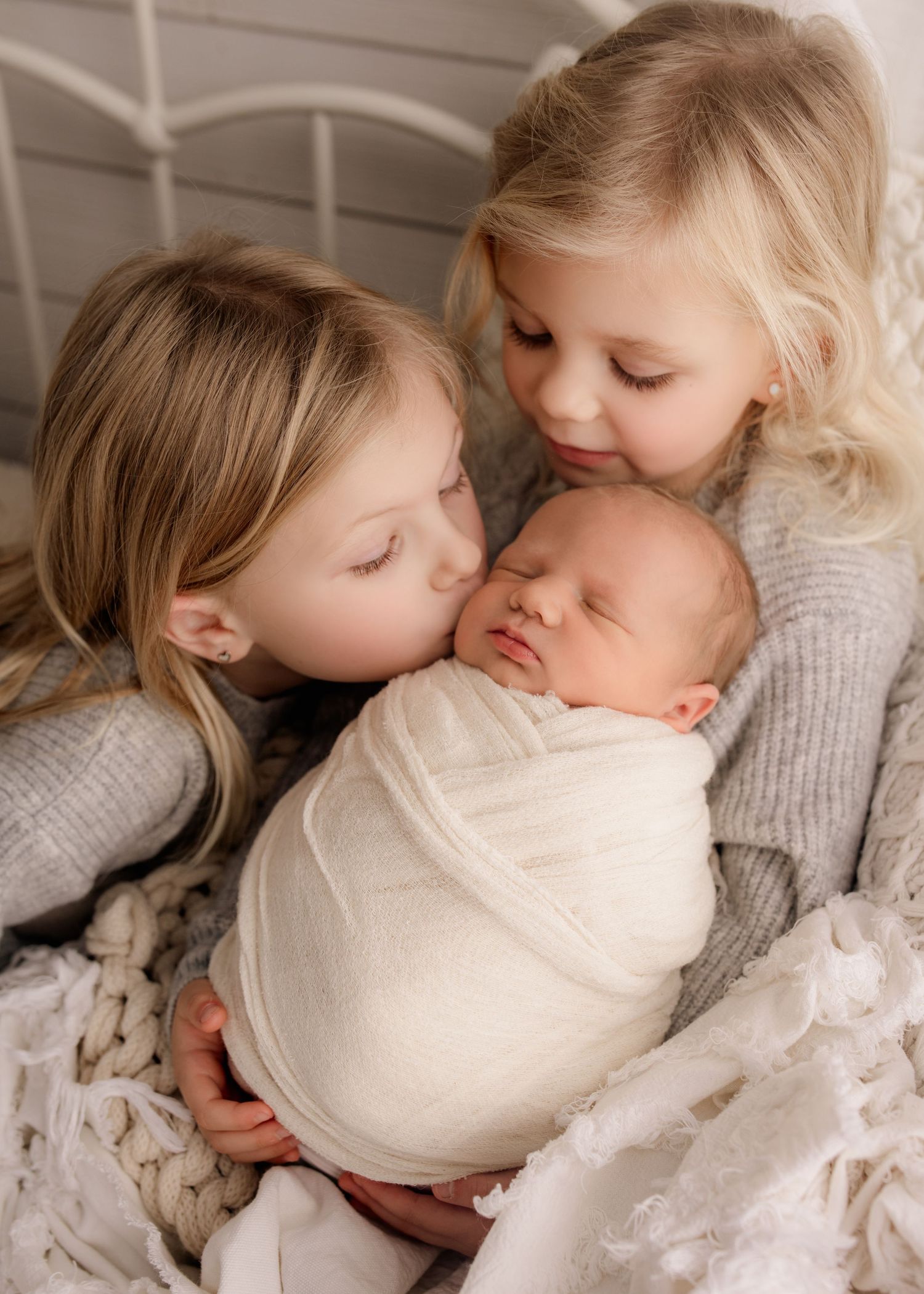 Two young sisters lovingly kissing and cuddling their swaddled newborn sibling on a cozy bed.