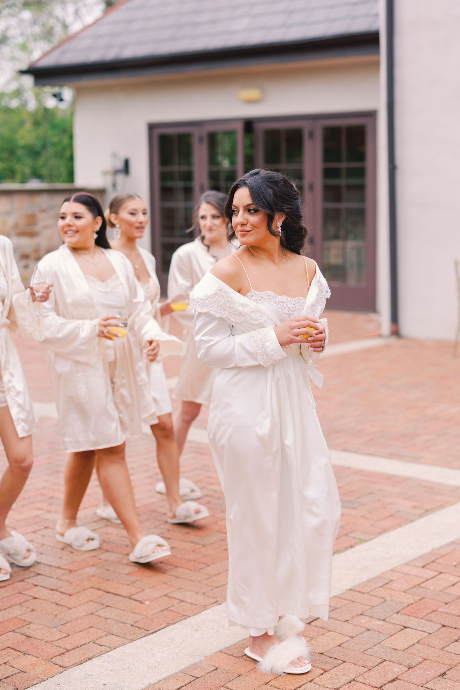Bride in white robe walks with bridesmaids holding mimosas on brick patio before wedding ceremony.