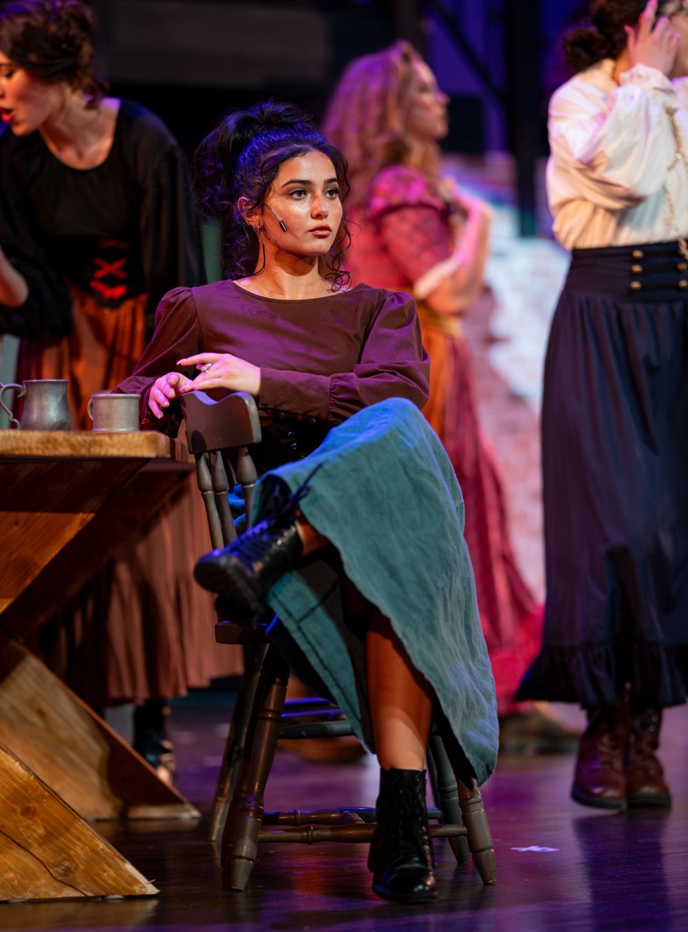 Performer in purple dress and turquoise skirt sits casually on wooden chair during theatrical production.