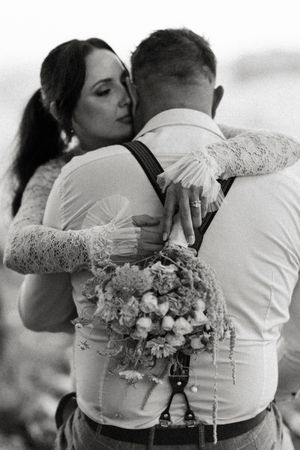 Black and white wedding photo of bride embracing groom from behind, holding a floral bouquet, wearing lace dress.