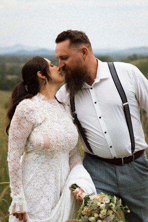 Bride in lace dress and groom with beard and suspenders share a kiss in a scenic outdoor field.