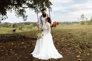 Bride and groom share a romantic moment by a tree swing on a rustic farm, bride holding a white bouquet.