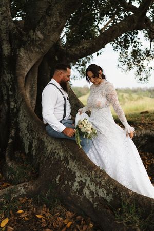 Bride in lace gown and groom in suspenders sit together in a large tree, sharing an intimate moment outdoors.