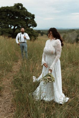Bride in white lace gown holds bouquet in field as groom in suspenders approaches smiling on their wedding day.
