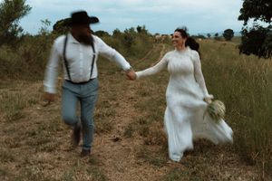 A bride in a white lace gown and groom in suspenders run hand-in-hand through a rustic field on their wedding day.